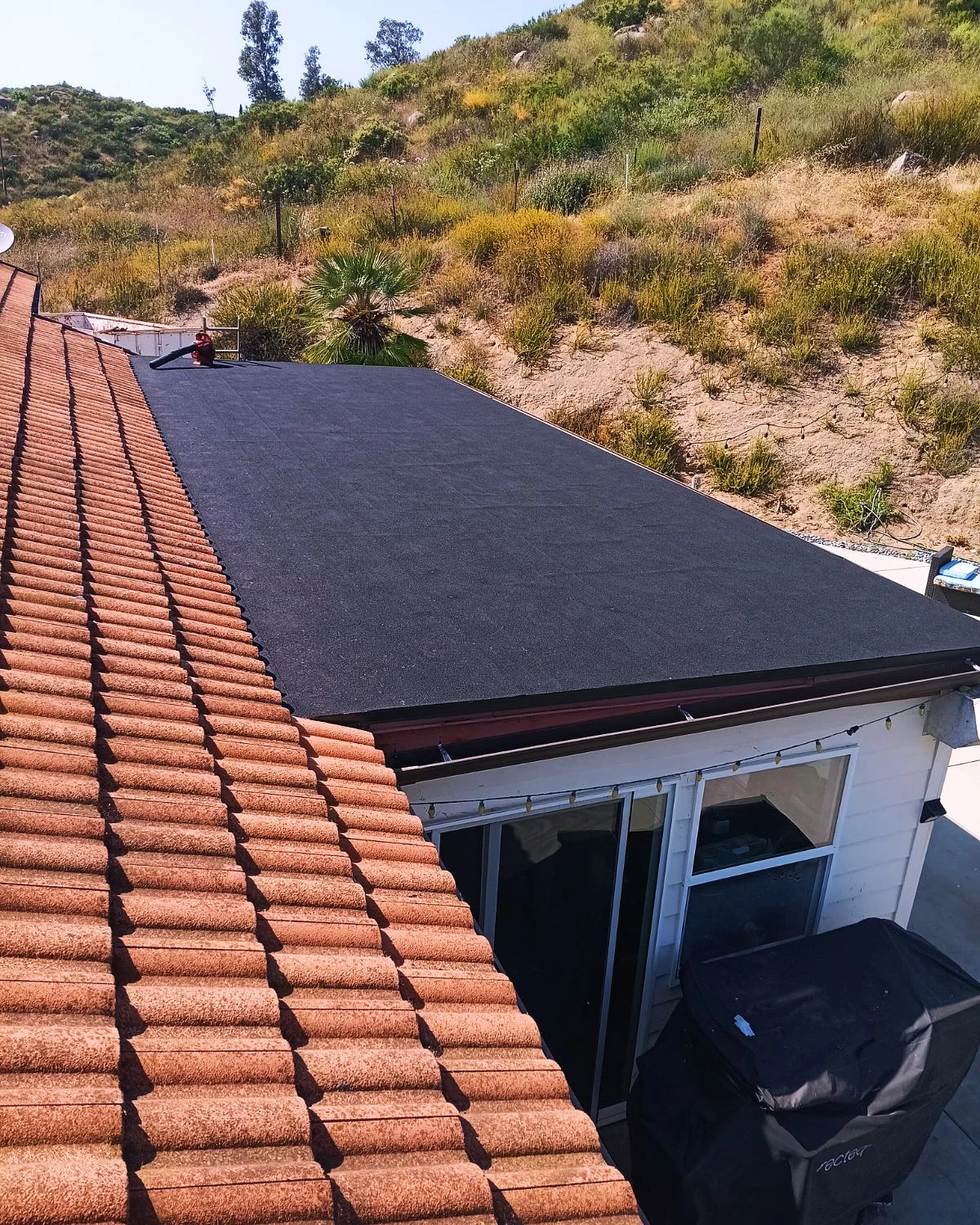 Dark asphalt flat roof installation on hillside home with white trim and clay tile roof visible